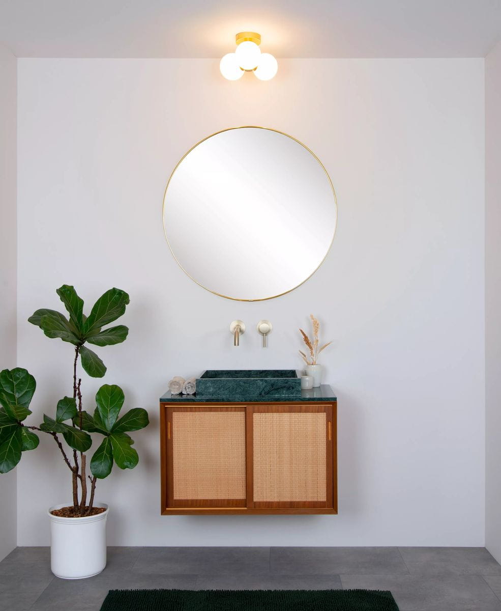 Lampa sufitowa Trudy 3-punktowa w kolorze złotym z trzema okrągłymi kloszami w białym kolorze. Miejsce montażu na gładkiej, jasnej ścianie, otoczenie z lustrem w złotej ramie i minimalistycznym umeblowaniem w łazience.