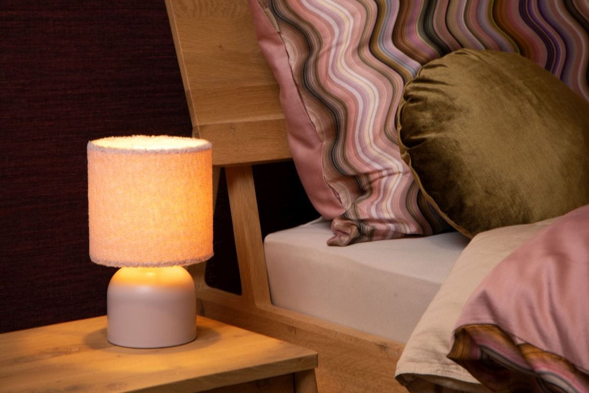 Różowa lampa stołowa Woolly z tkaninowym kloszem i okrągłą podstawą, emitująca ciepłe światło. Ustawiona na drewnianym stoliku obok łóżka, w otoczeniu poduszek w różnych odcieniach, na tle ciemnej ściany.