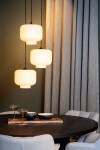 Lampa wisząca Medine 3-punktowa w kolorze czarnym i białym z trzema szklanymi kloszami w kształcie cylindrów. Ujęcie frontalne, zawieszona nad okrągłym stołem, otoczona szarymi zasłonami i akcesoriami stołowymi.