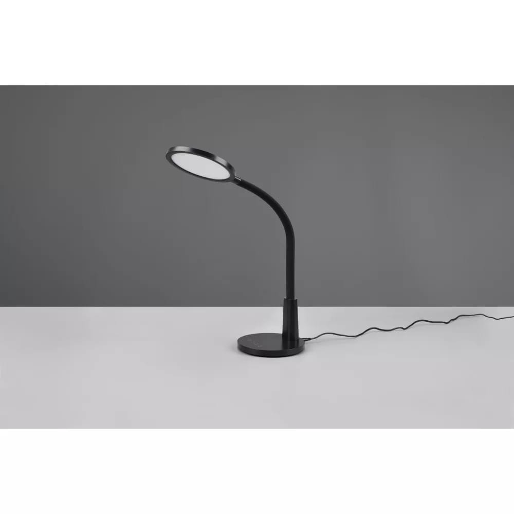 Czarna lampa biurkowa LED Sally z jednym punktem świetlnym, z elastycznym ramieniem i okrągłą podstawą. Ujęcie z boku na stole, z widocznym kablem zasilającym. Tło w odcieniu szarości.
