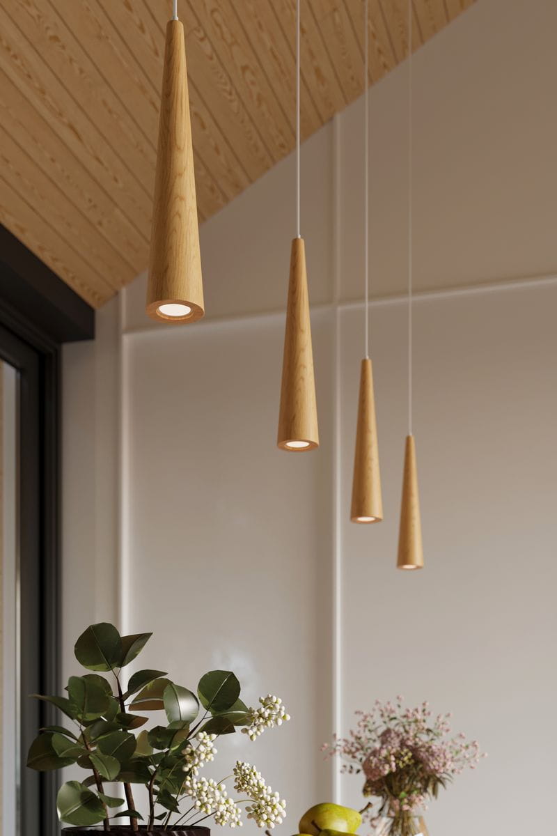 Lampa wisząca Sula 1-punktowa w kształcie stożka, wykonana z naturalnego drewna w jasnym odcieniu. Zawieszona na cienkim przewodzie, z widocznym źródłem światła u dołu. W tle dekoracyjne rośliny w doniczkach oraz jasne, minimalistyczne wnętrze.