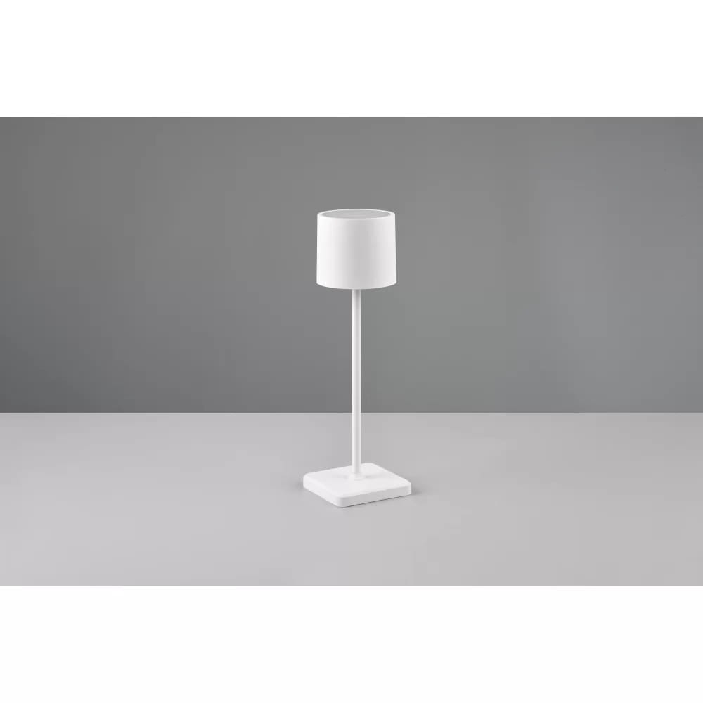 Biała lampa stołowa zewnętrzna Fernandez LED, z cylindrycznym kloszem i prostą, metalową podstawą, stojąca na szarej powierzchni. Klosz ma gładką fakturę, a całość prezentuje minimalistyczny styl.