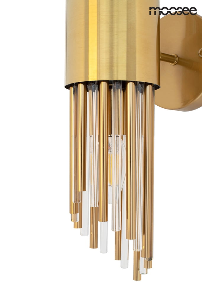 Kinkiet Amadeo 2-punktowy w kolorze złotym, z metalową podstawą i szklanymi prętami. Zbliżenie na detale, ukazujące błyszczące elementy oraz przezroczyste rurki, które tworzą elegancki wygląd. Produkt zamontowany na ścianie.