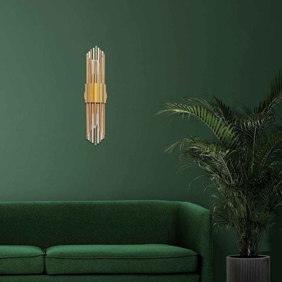 Kinkiet Amadeo 2-punktowy w złotym kolorze, złożony z prostokątnych elementów, zamontowany na ciemnozielonej ścianie. W tle widoczna zielona sofa i roślina doniczkowa, co podkreśla nowoczesny styl aranżacji wnętrza.