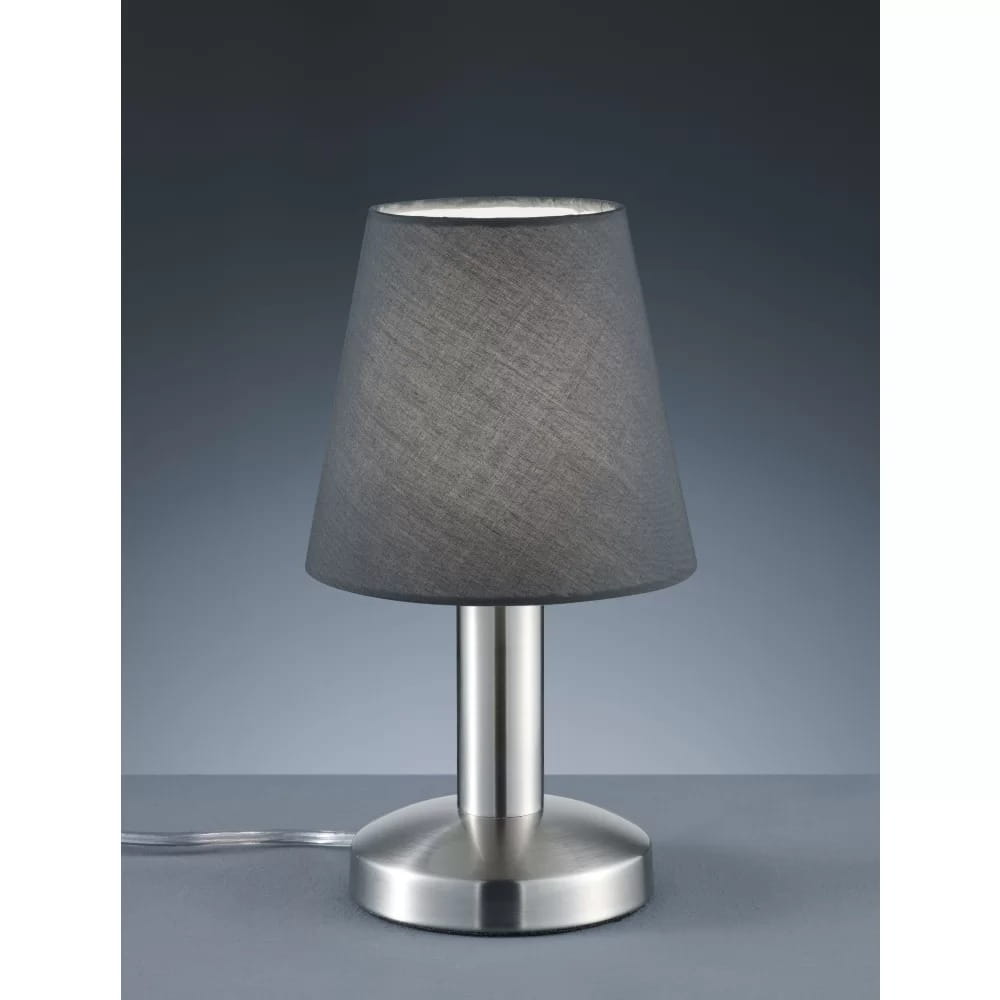 Lampa stołowa Mats II z podstawą z niklu i szarym kloszem z materiału, o stożkowym kształcie. Widok z przodu, na stole, z widocznym przewodem zasilającym. Prosty, elegancki design, idealna do użytku w różnych wnętrzach.