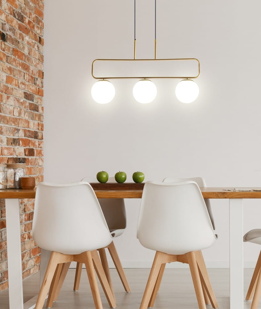 Lampa wisząca Jove 3-punktowa w kolorze mosiądzu z trzema okrągłymi kloszami. Zawieszona poziomo nad stołem, w nowoczesnej aranżacji z białymi krzesłami i drewnianym blatem. Tło z cegły podkreśla jej elegancki design.
