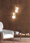 Lampa wisząca Cordel 4-punktowa w kolorze mosiężnym, z czterema białymi, okrągłymi kloszami. Zawieszona pionowo, na tle brązowej ściany. Widoczny również biały fotel i drewniany stół z książkami w aranżacji wnętrza.