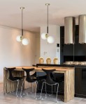 Duo lampa wisząca 2-punktowa w kolorze czarnym i złotym, z białymi szklanymi kloszami, zawieszona na długich kablach. Produkt umieszczony w nowoczesnej kuchni, nad drewnianym blatem z metalowymi krzesłami.