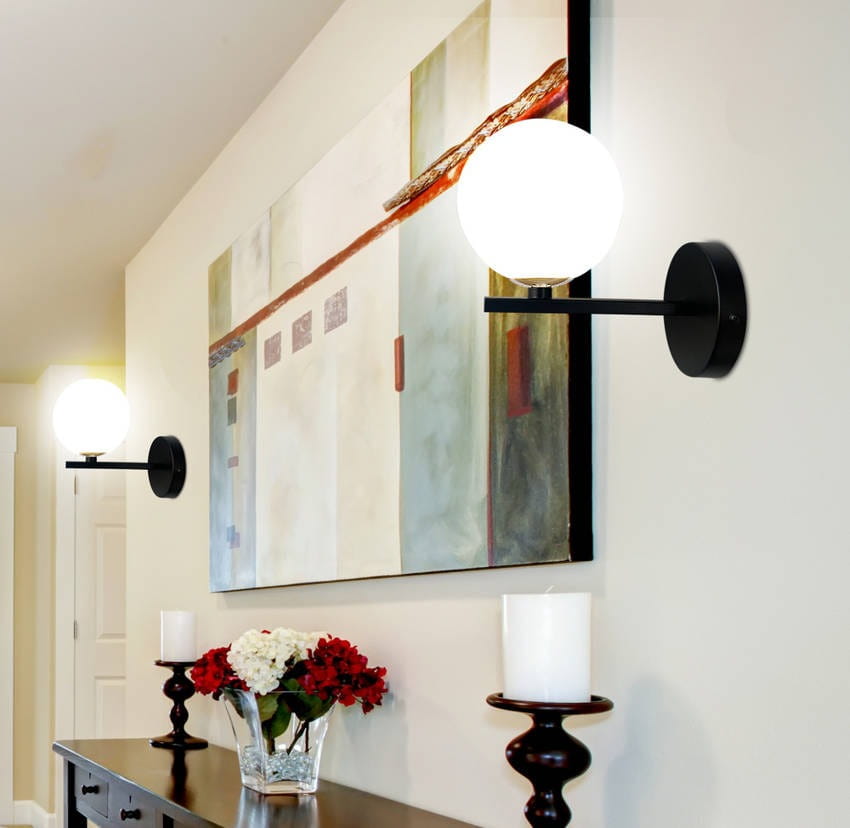 Kinkiet Kama 1-punktowy w kolorze czarnym i złotym z okrągłą, białą kloszową lampą. Ujęcie frontalne, zamontowany na ścianie obok dzieła sztuki. W tle widoczne kwiaty w wazonie i świeczniki.