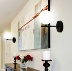 Kinkiet Kama 1-punktowy w kolorze czarnym i złotym z okrągłą, białą kloszową lampą. Ujęcie frontalne, zamontowany na ścianie obok dzieła sztuki. W tle widoczne kwiaty w wazonie i świeczniki.