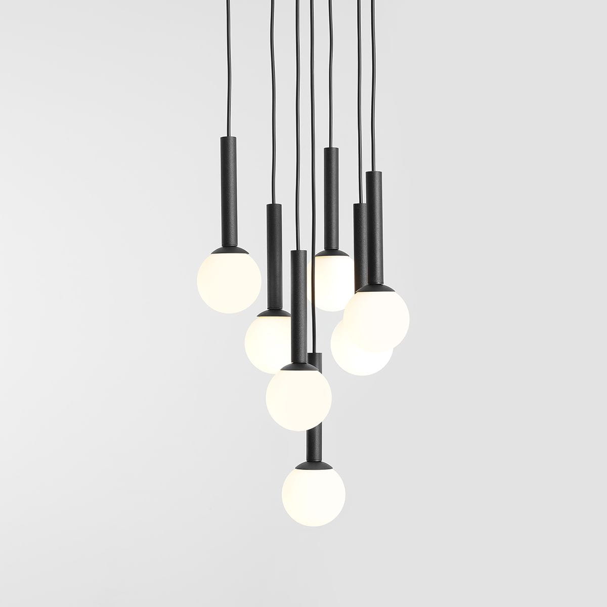 Lampa wisząca Riu 7-punktowa w kolorze czarnym, z siedmioma okrągłymi, białymi kloszami. Ujęcie z góry, pokazujące różne długości zwisających przewodów. Prosty, nowoczesny design, idealna do wnętrz. Tło jasnoszare.