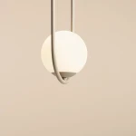 Lampa wisząca Riva 1-punktowa w kolorze beżowym z białym, okrągłym kloszem. Ujęcie z góry, lampa zawieszona na cienkim, metalowym stelażu, z wyraźnie widocznym elementem mocującym. Tło w jasnym odcieniu.