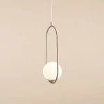 Lampa wisząca Riva 1-punktowa w kolorze beżowym, z okrągłym kloszem w białym kolorze, zawieszona na cienkim, białym przewodzie. Produkt ma minimalistyczny kształt z eliptycznym ramieniem, umieszczonym w centralnej części. Tło w odcieniu beżowym.