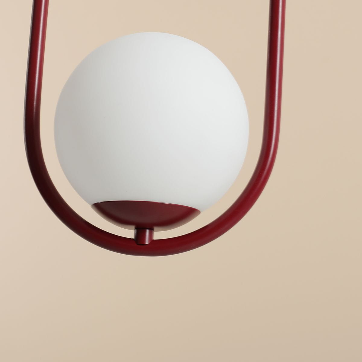 Lampa wisząca Riva 1-punktowa w kolorze czerwonym, z białą, okrągłą kloszem. Posiada metalowy, czerwony uchwyt w formie półokręgu. Ujęcie z dołu, ukazujące detal konstrukcji i elegancką formę. Tło w jasnym odcieniu.