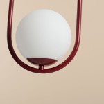 Lampa wisząca Riva 1-punktowa w kolorze czerwonym, z białą, okrągłą kloszem. Posiada metalowy, czerwony uchwyt w formie półokręgu. Ujęcie z dołu, ukazujące detal konstrukcji i elegancką formę. Tło w jasnym odcieniu.