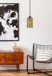 Cox lampa wisząca 1-punktowa w kolorze czarnym i złotym, cylindryczna, zawieszona na czarnym kablu. W tle widoczna mapa na ścianie oraz drewniana komoda z książkami i dekoracjami, w otoczeniu nowoczesnego czarnego fotela.