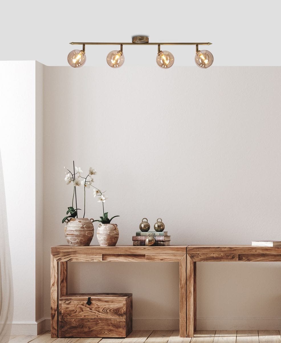 Lampa sufitowa Rodia 4-punktowa w kolorze patyny i bursztynowym, montowana na suficie. Posiada cztery okrągłe klosze, które emitują ciepłe światło, umieszczone na metalowej ramie. W tle widoczna drewniana konsola z roślinami i dekoracjami.