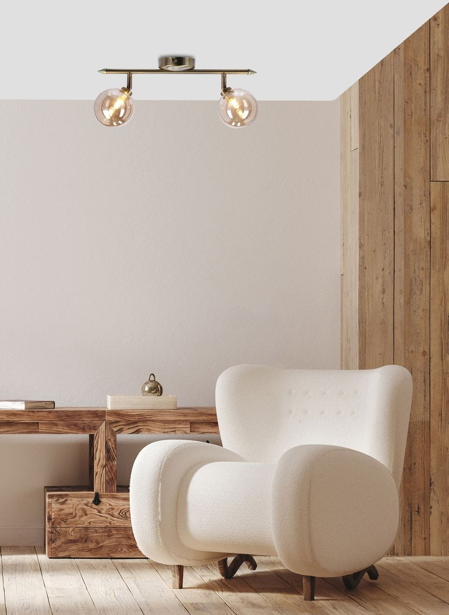 Lampa sufitowa Rodia 2-punktowa w kolorze patyny z bursztynowymi kloszami. Produkt montowany na suficie, z widocznymi żarówkami, zawieszony na metalowej konstrukcji z dwiema ramionami. Tło w jasnych odcieniach, w aranżacji z nowoczesnym fotelem.