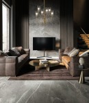 Lampa wisząca Arco 6-punktowa w mosiężnym wykończeniu z przezroczystymi kloszami. Zawieszona centralnie w nowoczesnym wnętrzu, w towarzystwie brązowych sof i okrągłego stolika kawowego. Tło z szarego kamienia dodaje elegancji.