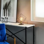 Lampa stołowa Alambre 1-punktowa z czarną metalową ramą w kształcie diamentu, złotą oprawą żarówki i szklaną kulą. Ustawiona na białym stole obok świeczników i niebieskiego fotela. Tło w odcieniach szarości.