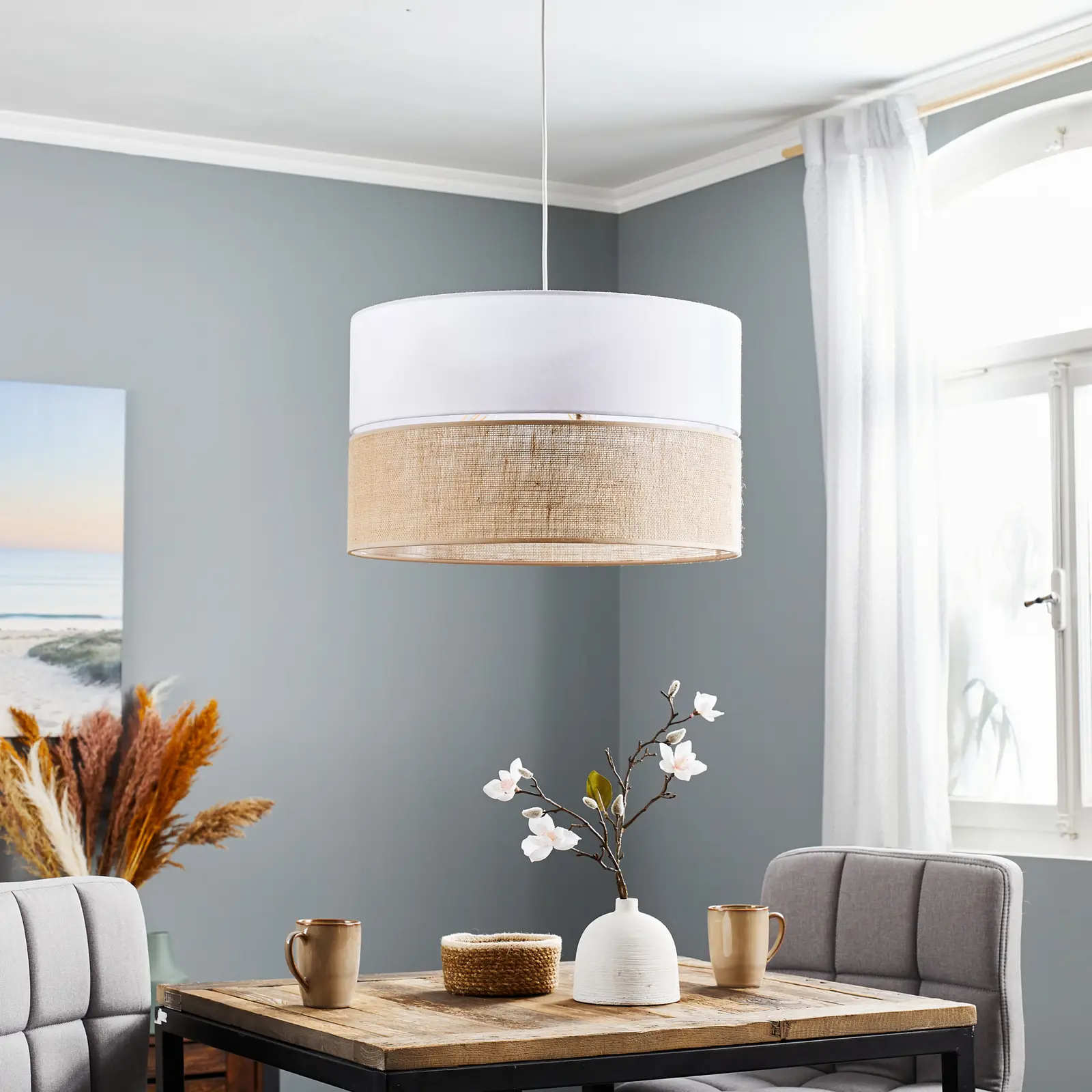 Lampa wisząca Linobianco 3-punktowa w kolorze białym i jutowym, o cylindrycznym kształcie, zawieszona nad drewnianym stołem. W tle widoczne szare ściany, zasłony, roślinność i dekoracje.