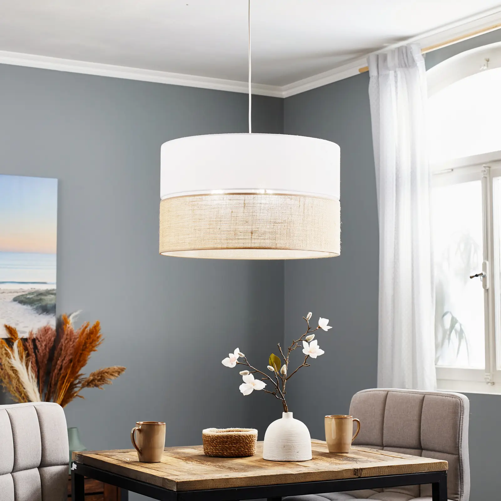 Lampa wisząca Linobianco 3-punktowa w kolorze białym z jutowym detalem, zawieszona nad drewnianym stołem w jadalni. Otoczona jasnymi krzesłami, dekoracyjnym wazonem z kwiatami i filiżankami, na tle szarej ściany i okna z firankami.