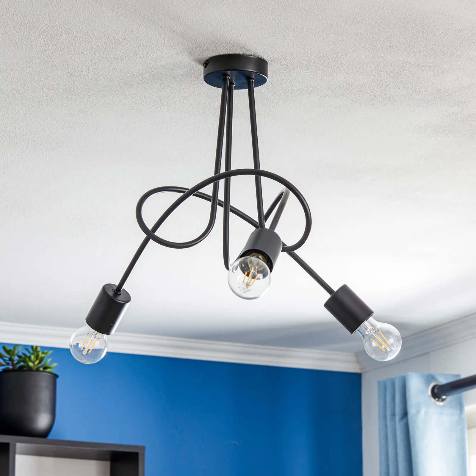 Lampa wisząca Tango 3-punktowa w kolorze czarnym z trzema żarówkami w przezroczystych oprawach. Kable tworzą dynamiczny wzór, mocowane do sufitu. Tło w niebieskim odcieniu, z rośliną w doniczce w tle.