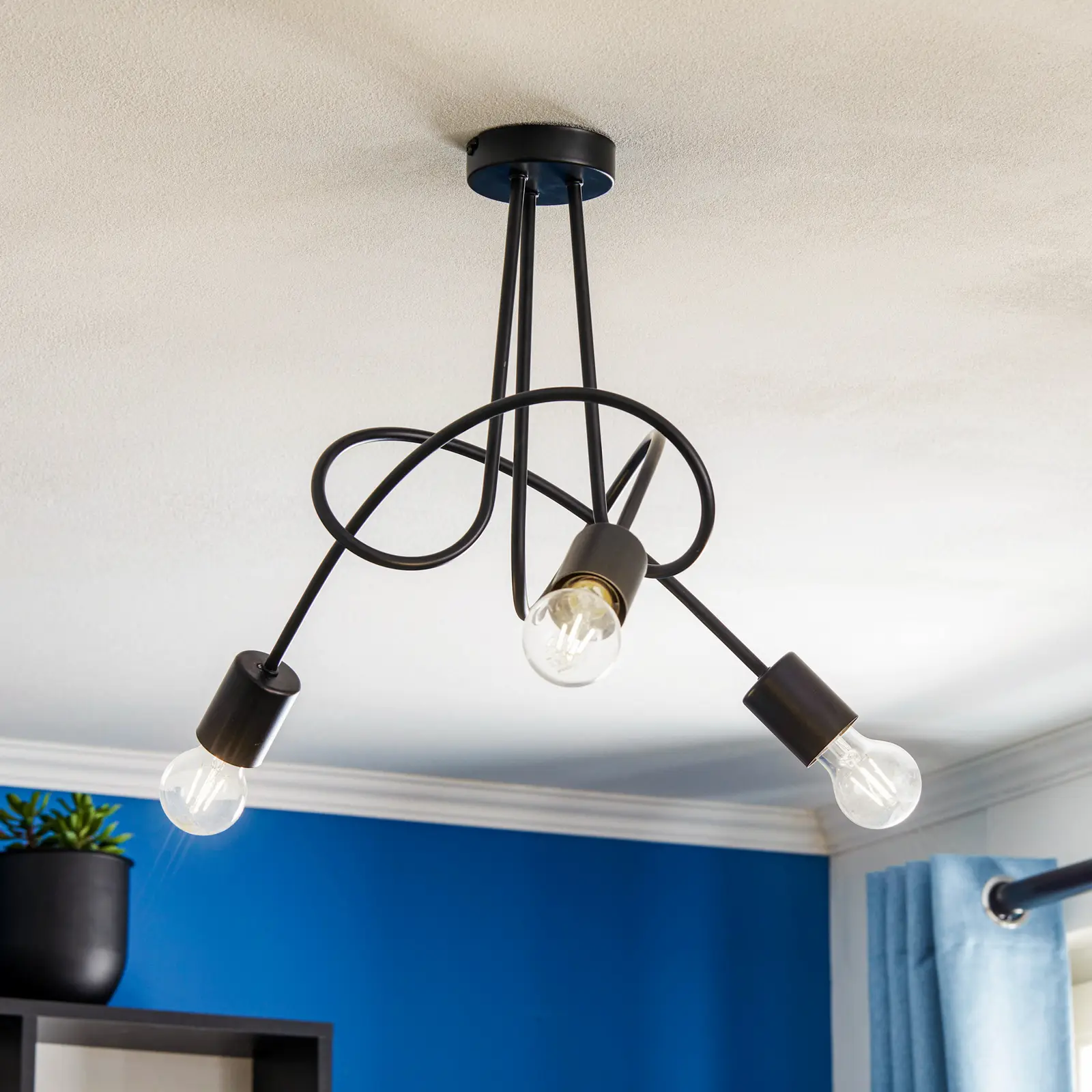 Czarna lampa wisząca Tango z trzema punktami świetlnymi, z metalowymi ramionami w kształcie spirali. Lampa zamontowana na suficie, z widocznymi żarówkami o przezroczystych kloszach. Tło w kolorze niebieskim, uzupełnione rośliną w doniczce.