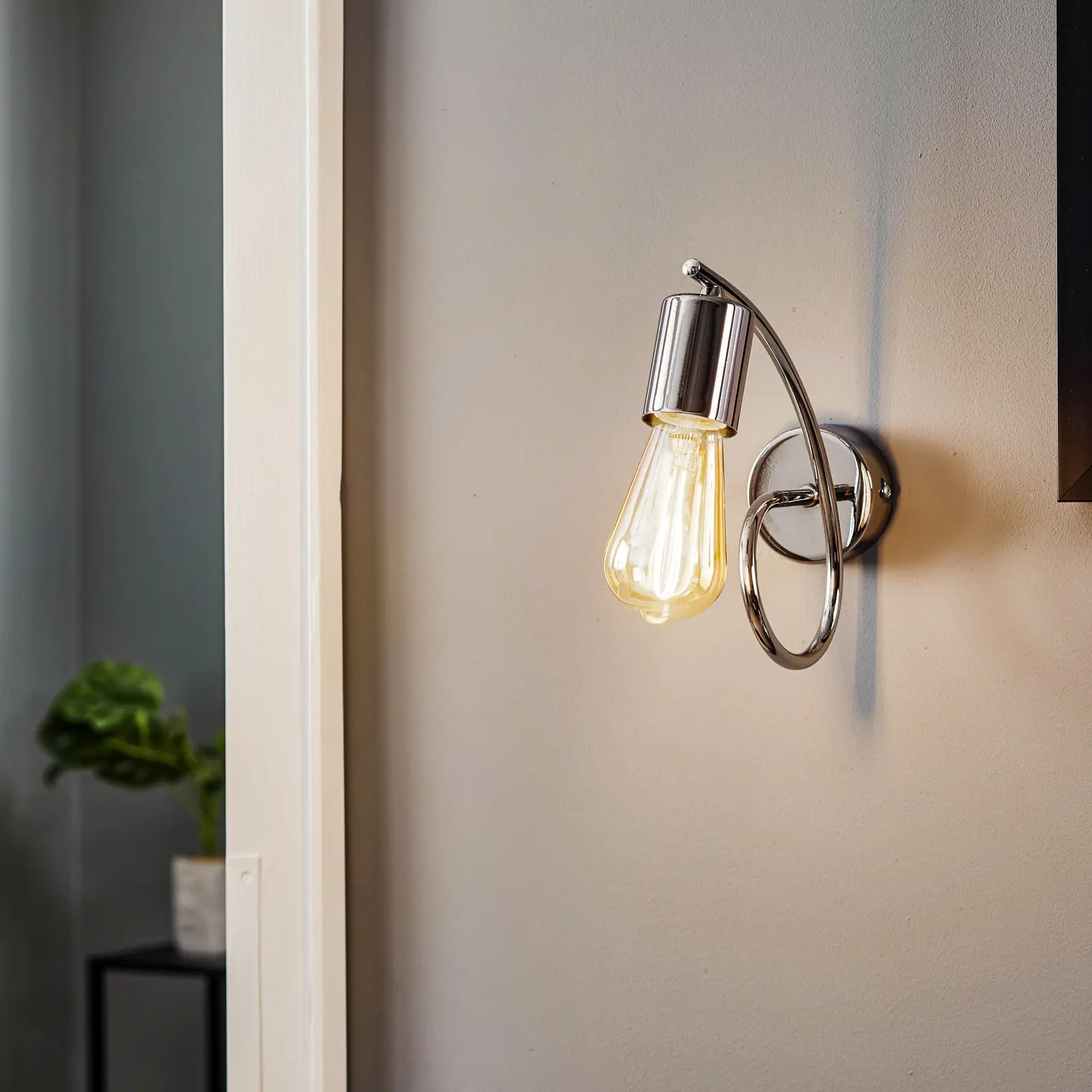 Kinkiet Tango 1-punktowy w chromie, z okrągłą podstawą i elastycznym ramieniem, prezentujący żarówkę w kształcie kropli. Zawieszony na ścianie, tworzy nowoczesny akcent w aranżacji wnętrza. Tło w neutralnym kolorze.