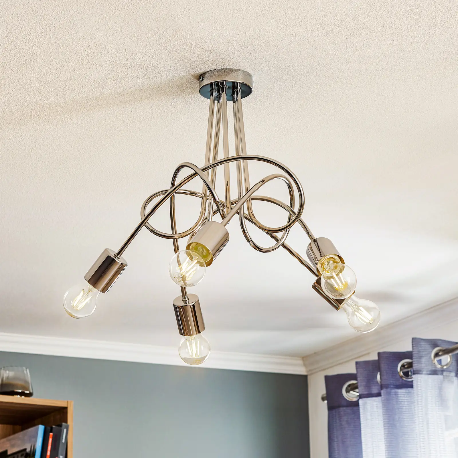 Lampa wisząca Tango 5-punktowa o chromowanej konstrukcji, z pięcioma żarówkami w szklanych kloszach. Ujęcie z góry, zamontowana na suficie, z dekoracyjnymi, krętymi ramionami. Tło w stonowanych kolorach.