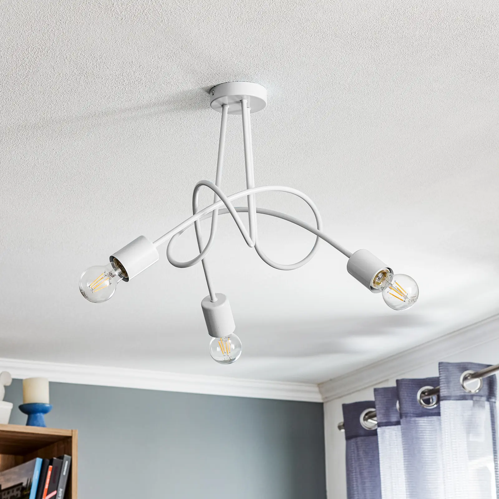 Lampa wisząca Tango 3-punktowa w kolorze białym, z trzema żarówkami na skręconych przewodach. Ujęcie z dołu, wisząca pod sufitem. W tle szare ściany oraz zasłony. Prosta konstrukcja z estetycznymi detalami.