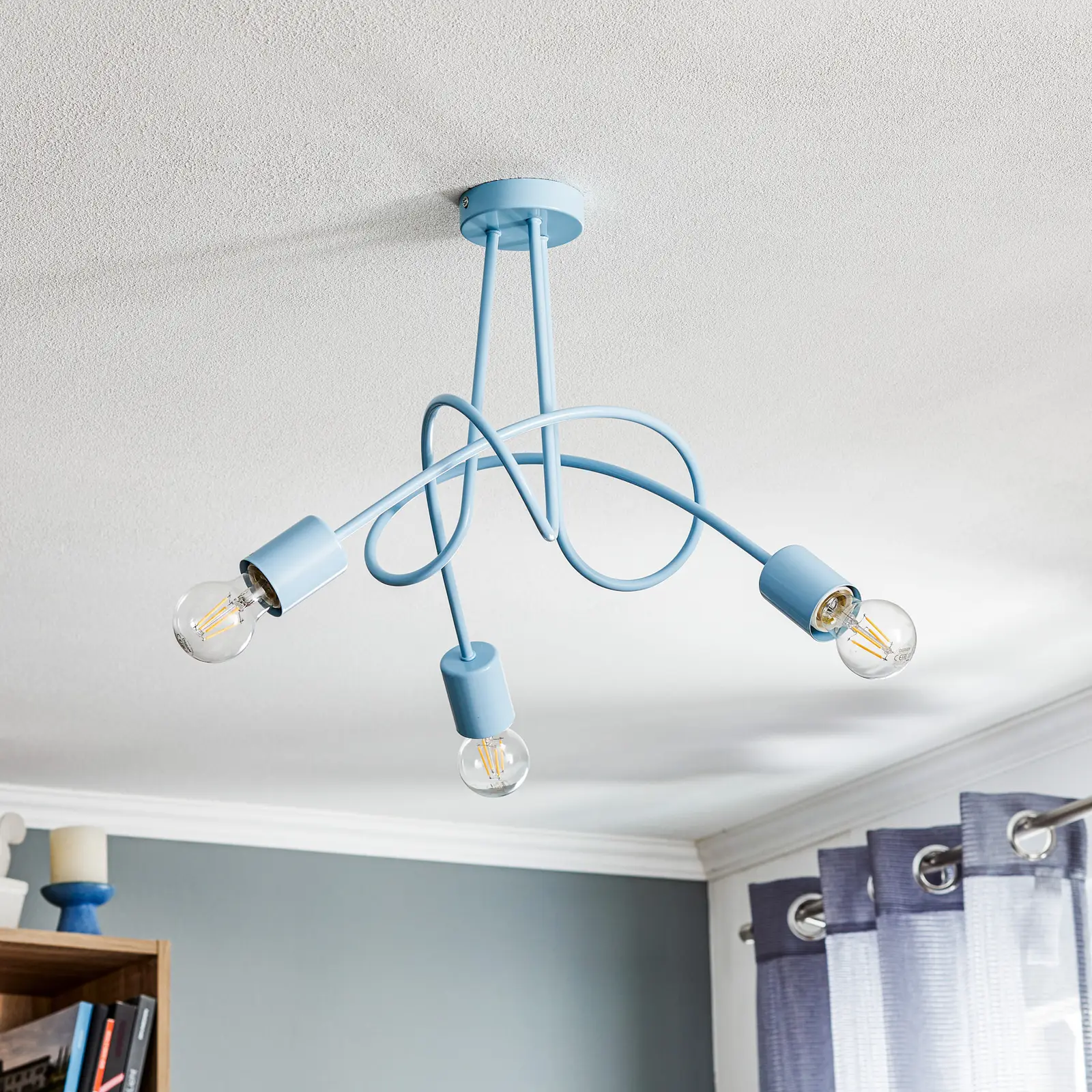 Lampa wisząca 3-punktowa Tango w kolorze niebieskim z trzema zakrzywionymi ramionami. Kable w pastelowym odcieniu prowadzą do opraw z przezroczystymi żarówkami, zamontowana na sufitowym mocowaniu.