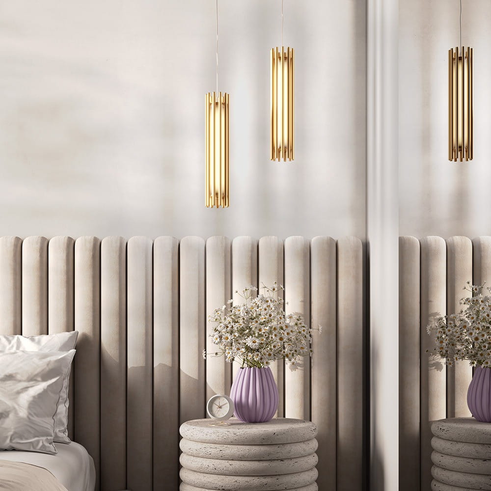 Lampa wisząca Sonata LED, 1-punktowa, wykonana z mosiądzu, o cylindrycznym kształcie, zawieszona nad stolikiem nocnym. W tle widoczna dekoracyjna ściana z pionowymi panelami oraz wazon z kwiatami. Wnętrze w jasnych odcieniach.