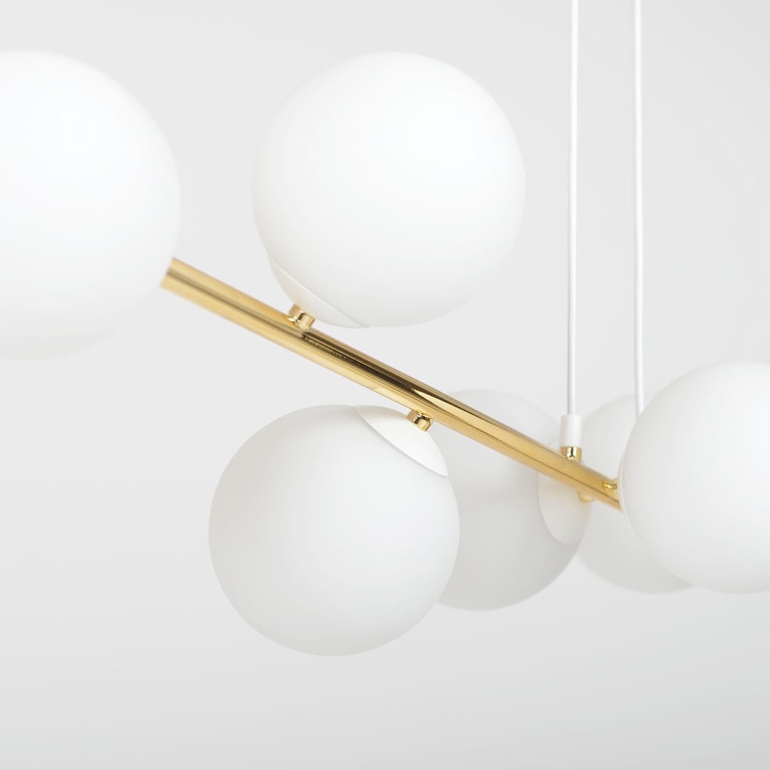Lampa wisząca Gravity 6-punktowa w kolorze białym i złotym. Posiada sześć opalowych kul, zawieszonych na złotej metalowej belce. Ujęcie z bliska, ukazujące detale konstrukcji i materiałów. Tło jasne.