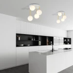 Lampa sufitowa Dolce 4-punktowa w kolorze białym i złotym, z opalowymi kloszami, zawieszona na długich złotych linkach. Widok z dołu, umieszczona w nowoczesnej kuchni z białymi i czarnymi meblami.