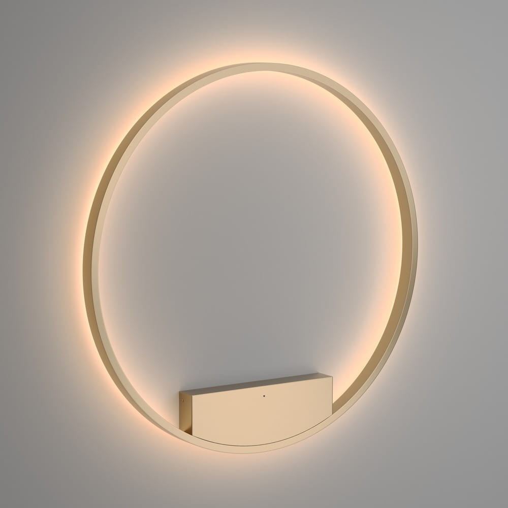 Kinkiet Rim D80 LED w kolorze mosiądzu, o okrągłym kształcie. Zainstalowany na ścianie, emituje miękkie światło wokół obręczy. Dolna część z prostokątnym korpusem, w kolorze mosiądzu, widoczna z przodu.