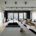 Czarna lampa sufitowa 1-punktowa, okrągła, zamontowana na suficie, emitująca światło w kierunku stołu w nowoczesnym wnętrzu. Widoczny jasny stół, zlew i strefa wypoczynkowa w tle. Czyste linie, minimalistyczny design.