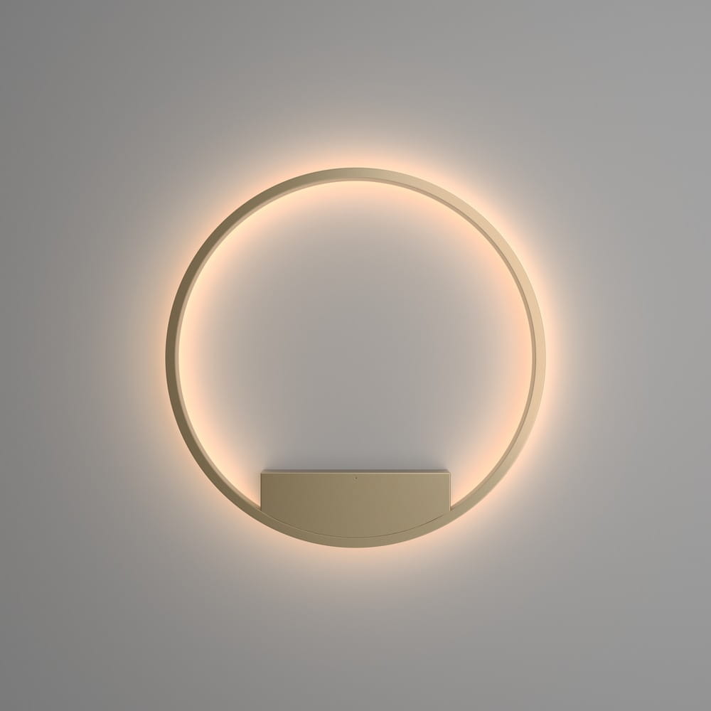 Kinkiet Rim D60 LED w mosiężnym kolorze, w kształcie okręgu, zamontowany na ścianie. Emituje miękkie, ciepłe światło wokół obwodu. Posiada prostą, minimalistyczną formę z płaskim, dyskretnym mocowaniem. Tło w jasnym odcieniu.