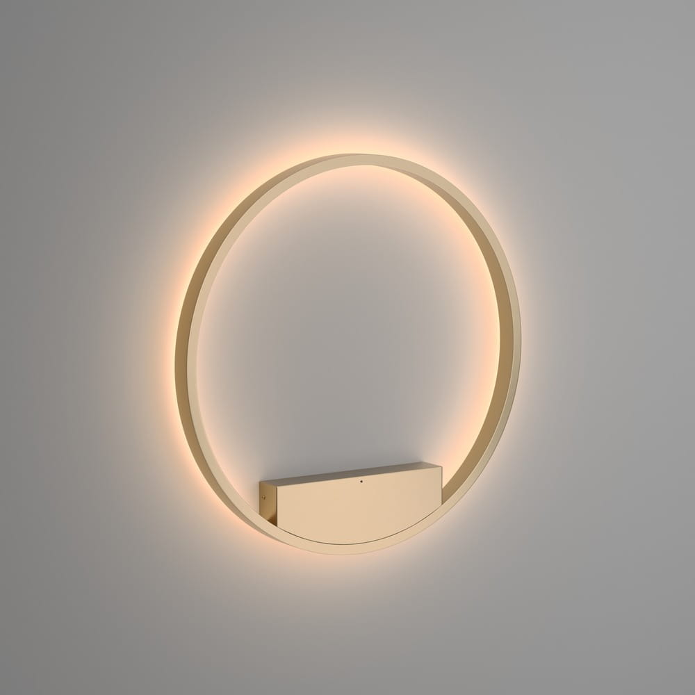 Kinkiet Rim D60 LED w kolorze mosiądzu, o okrągłym kształcie. Element montażowy w formie prostokąta znajduje się w dolnej części. Oświetlenie emituje delikatne światło, tworząc efekt halo na ścianie. Produkt zamontowany na jasnym tle.