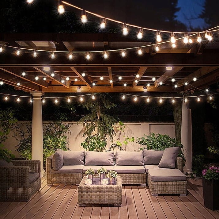 Girlanda ogrodowa o długości 15 m w kolorze czarnym z 15 żarówkami LED E27 1W, zawieszona nad zestawem mebli ogrodowych. Żarówki emitują ciepłe światło, tworząc przyjemną atmosferę w otoczeniu roślinności.