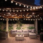 Girlanda ogrodowa o długości 15 m w kolorze czarnym z 15 żarówkami LED E27 1W, zawieszona nad zestawem mebli ogrodowych. Żarówki emitują ciepłe światło, tworząc przyjemną atmosferę w otoczeniu roślinności.