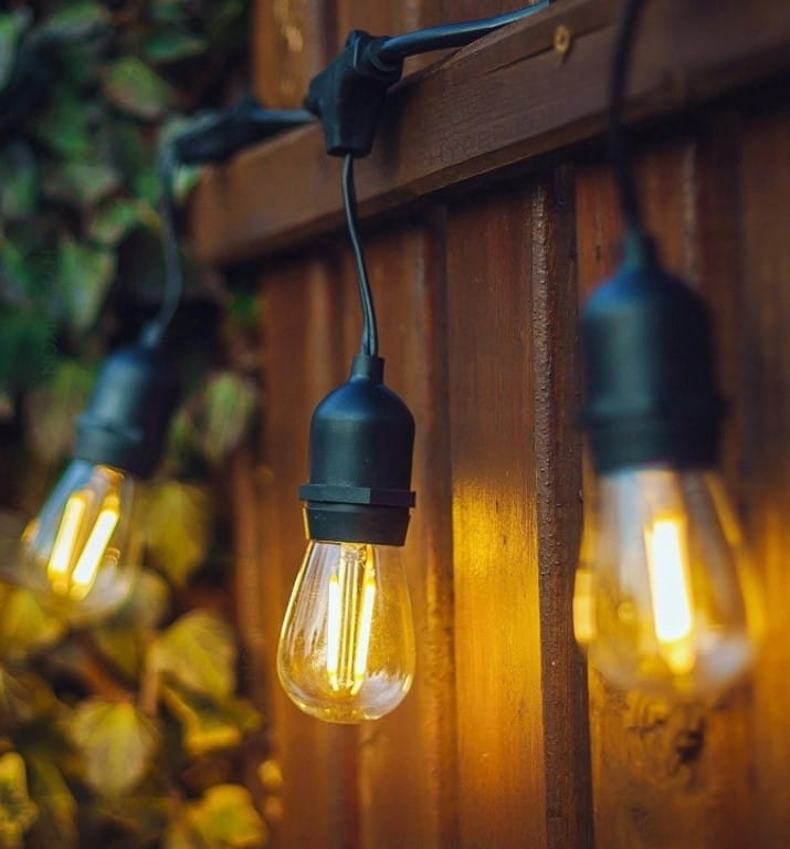 Girlanda ogrodowa o długości 15 m z czarnym przewodem, zawierająca 15 żarówek LED E27 1W. Żarówki w stylu retro, wiszące na drewnianym tle, emitujące ciepłe światło. Kąt ujęcia zbliżenie na detale żarówek.