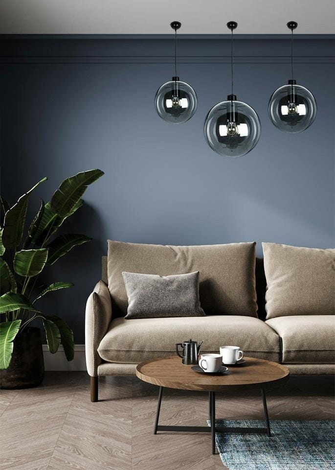 Lampa wisząca Sandra 30, czarna, o okrągłym kształcie, zawieszona na cienkim kablu. W tle widoczny stylowy salon z jasnoniebieską ścianą, brązową sofą i okrągłym stolikiem kawowym. Dodatkowo roślina w doniczce po lewej stronie.