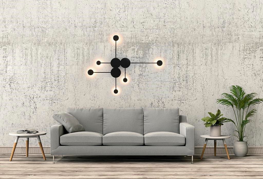 Kinkiet Shadow Multi LED 6-punktowy w kolorze czarnym, zamontowany na ścianie, składający się z okrągłych źródeł światła oraz prostych ramion. W tle widoczna szara sofa, stół oraz roślinność, na złożonym tle z betonu.
