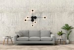 Kinkiet Shadow Multi LED 6-punktowy w kolorze czarnym, zamontowany na ścianie, składający się z okrągłych źródeł światła oraz prostych ramion. W tle widoczna szara sofa, stół oraz roślinność, na złożonym tle z betonu.