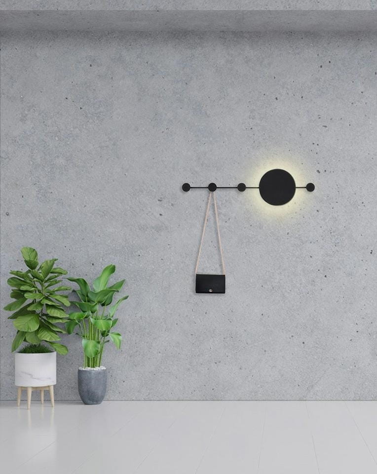 Kinkiet Shadow Hang LED w kolorze czarnym, z okrągłym źródłem światła i wieszakiem na torbę. Zamontowany na szarej ścianie, w towarzystwie roślin doniczkowych. Prosta, nowoczesna forma, idealna do wnętrz.