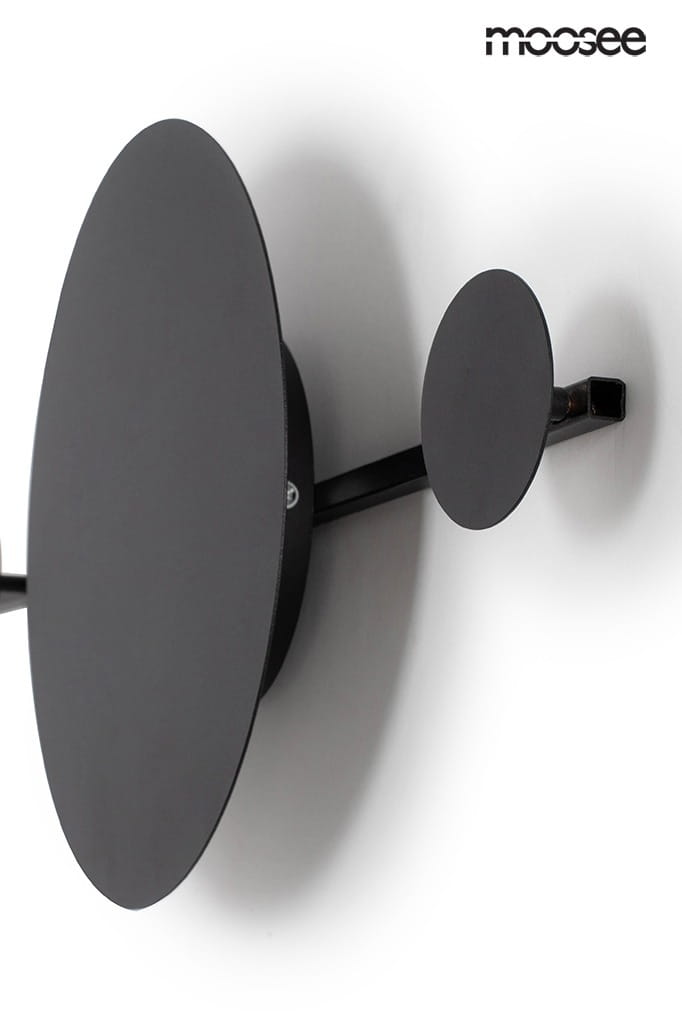 Czarny kinkiet LED Shadow Hang z wieszakiem, składający się z dużej owalnej oraz małej okrągłej części. Produkt zamontowany na ścianie, w minimalistycznym stylu, z widocznym metalowym mocowaniem. Tło jest jasne, co podkreśla ciemny kolor lampy.