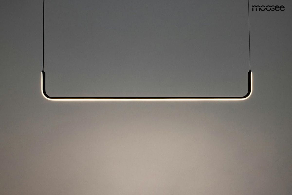 Czarna lampa wisząca Shape 90 LED w formie prostokątnej, zawieszona na cienkich linkach. Emituje ciepłe światło z subtelnym podświetleniem krawędzi. Tło w odcieniach szarości, co podkreśla minimalistyczny design.