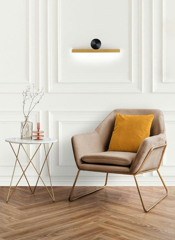 Kinkiet Wand 45 LED w kolorze złotym, z czarnym okrągłym elementem. Montowany na ścianie, z prostokątną, podświetlaną belką. W tle elegancki fotel z poduszką w kolorze musztardowym oraz biały stół z marmurowym blatem i złotymi nogami.