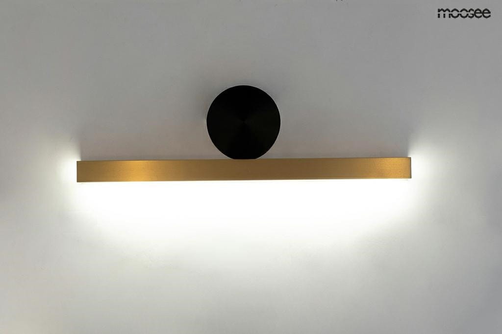Kinkiet Wand 45 LED w złotym kolorze, umieszczony na białej ścianie. Posiada prostokątną, matową oprawę oraz okrągły czarny element montażowy. Emituje ciepłe światło z obu końców, tworząc nowoczesny efekt oświetleniowy.